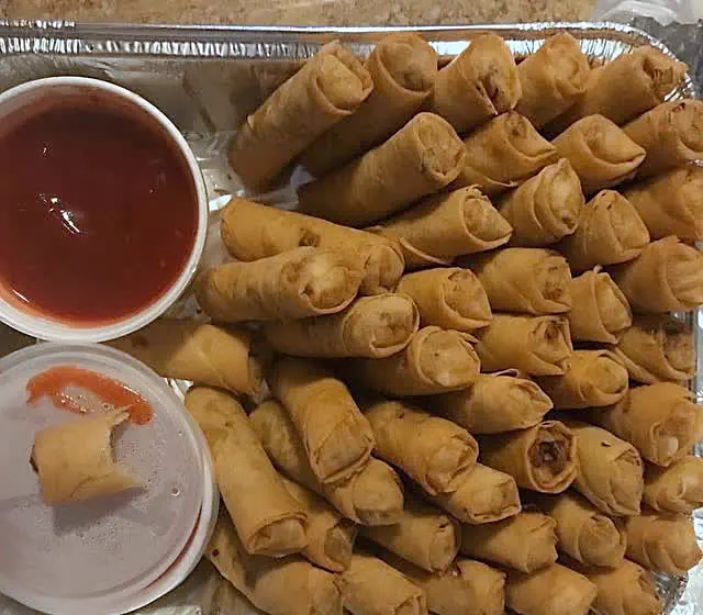 A foil party tray filled with lumpia, with cups of sweet chili sauce