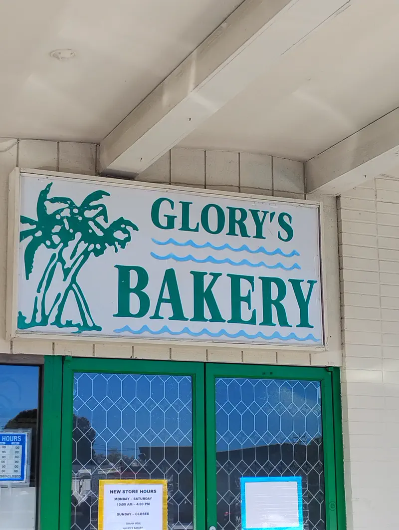 The painted Glory’s Bakery sign, with palm trees and blue waves, over the green doors of the Aragona Blvd shop